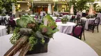 A basket of fresh vegetables are used as centerpieces on a room full of small round event tables.