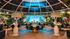 A blue-lit stage with tables, chairs, and oversized floor lamps bring life to an evening event under a glass dome.