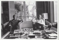 A black-and-white photo of two men in a living room, sitting behind a coffee table filled with books.