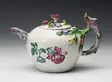 A photo of a teapot from 1750. A deep beet purple flower and green leaves/stem adorn the side. An ornate flower sits on top and its stem stretches downward as the teapot handle.