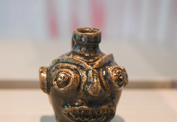 A tiny jug dark green and brown jug with a carved face sits on a table.