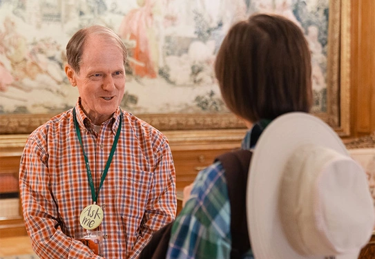 A person wearing an "Ask Me" badge talks to another person in front of a large hanging tapestry.