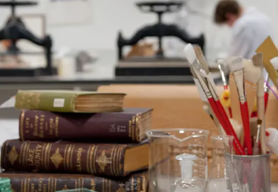 A table in a laboratory setting with stacked books, glass labware, and brushes. A person works in the background.