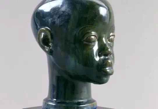 Three-quarter profile view of a small blue/green glazed terracotta sculpture of a young Black boy's head and neck.