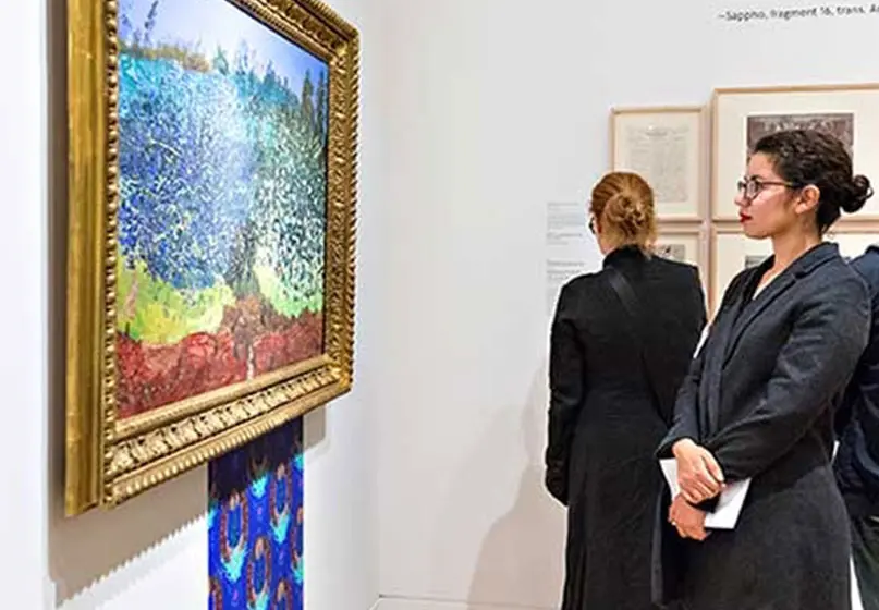 A woman in glasses and her arms folded in front of her holding some kind of white envelope or paper, looks at a blue, green, and red oil-drawn painting. What looks like a rectangular blue wallpaper swatch is attached to the wall below the painting.