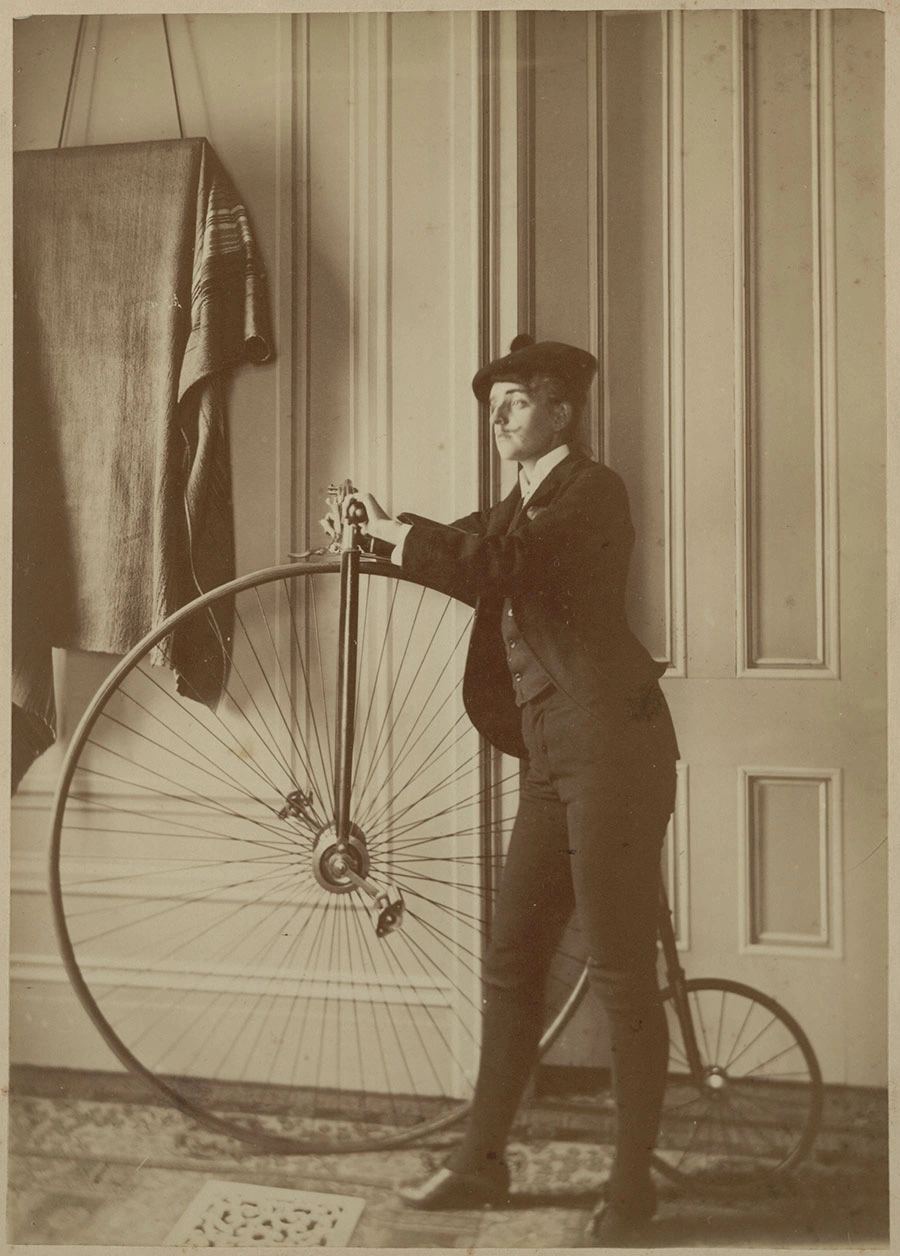 Black-and-white photo of Frances B. Johnston dressed in a suit, posing with a penny-farthing.