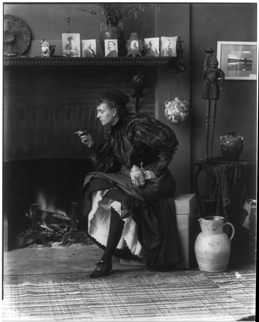 Black-and-white photo of Frances B. Johnston sitting in front of a fireplace, looking off-camera with a cigarette in her hand.