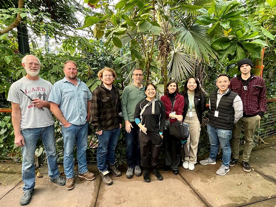 A group of people pose for a picture, standing in front of tropical trees.