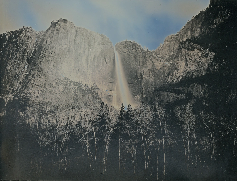 A waterfall cuts through a crevice in a tall mountain range, disappearing behind a forest of trees.