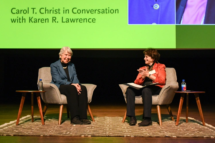 Two people sit in chairs on a stage. A large projection screen reads “Carol T. Christ in Conversation with Karen R. Lawrence.”